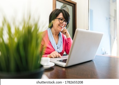 Sixty Year Old Female Teacher Wearing Headphones Having Online Class Via Video Chat On Laptop Computer. She Is Sitting On A Wooden Modern Desk At Home. Smiling And Enjoying Communication