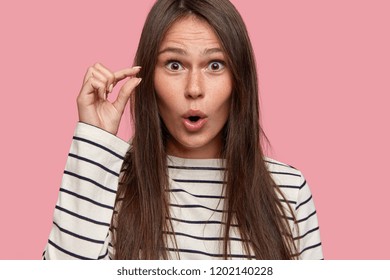 Size Concept. Close Up Shot Of Amazed Attractive Female Shapes Small Object With Hand, Demonstrates Tiny Thing, Dressed In Oversized Casual Sweater, Isolated Over Pink Background. It Costs Not Much