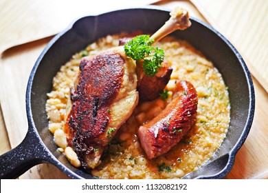Sizzling hot, rich, slow-cooked Trio Cassoulet of duck confit, pork belly and smoked sausage on a bed of white beans in a black cast iron pan on a wooden table.