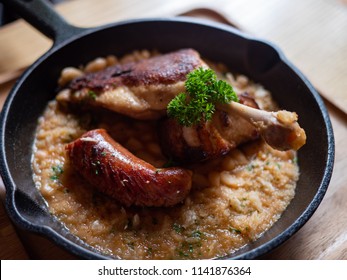Sizzling hot, rich, slow-cooked Trio Cassoulet of duck confit, pork belly and smoked sausage on a bed of white beans in a black cast iron pan on a wooden table. Natural Light.