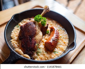 Sizzling hot, rich, slow-cooked Trio Cassoulet of duck confit, pork belly and smoked sausage on a bed of white beans in a black cast iron pan on a wooden table. Natural Light. - Powered by Shutterstock