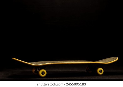 A skateboard is sitting on a black surface. The skateboard is old and worn, with yellow wheels. The image has a moody and somewhat melancholic feel, as the skateboard appears to be abandoned - Powered by Shutterstock