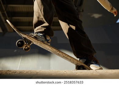 Skateboarder performing trick on concrete ramp in skatepark under large industrial roof casting shadows. Focus on skateboard and legs of skater wearing brown pants and blue sneakers - Powered by Shutterstock