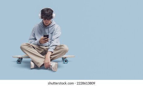 Skater wearing headphones and sitting on his skate, he is connecting with his smartphone and listening to music, blank copy space - Powered by Shutterstock