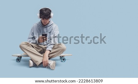 Skater wearing headphones and sitting on his skate, he is connecting with his smartphone and listening to music, blank copy space