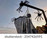 a skeleton in a black cloak hanging on a gallows. giant spiders descend on a thread towards him. outdoor arrangement in sunset.political satire