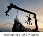 a skeleton in a black cloak hanging on a gallows. giant spiders descend on a thread towards him. outdoor arrangement in sunset.political satire