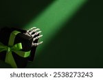 Skeleton hand emerging from black gift box with green ribbon on dark green background, mysterious Halloween present concept. Copyspace.