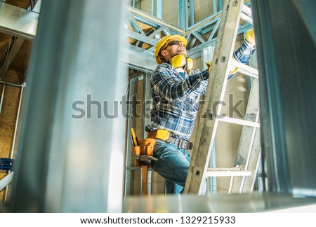 Skeleton Steel Building Construction Worker Wearing Safety Hard Hat and Noise Reduction Headphones. Going Up Using Ladder.