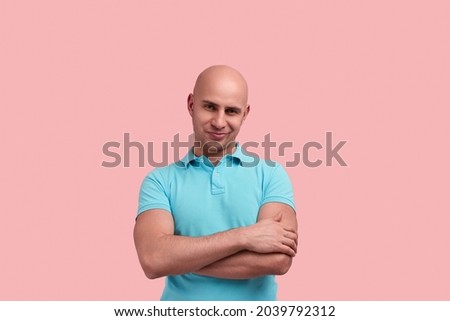 Skeptical bald homosexual man with bristle crosses hands over chest, does not believe information, makes wry mouth, inclines head aside, gay friendly, wears blue polo shirt, poses on pink background