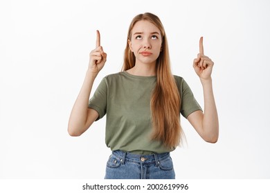 Skeptical Blond Girl Smirk, Pointing Fingers Up And Looking At Product Banner With Judging Displeased Face, Dislike Item Above, Standing Over White Background