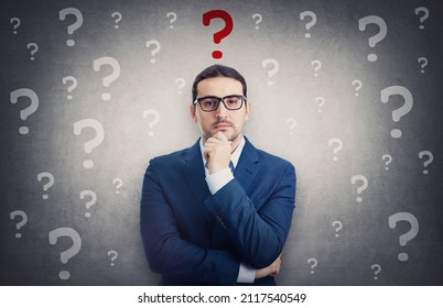 Skeptical businessman surrounded by interrogation marks has a main question as a red symbol over head. Thoughtful business person keeps hand under chin, waiting for an answer - Shutterstock ID 2117540549