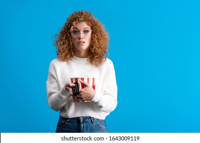 Skeptical Girl Watching Tv With Remote Control And Bucket Of Popcorn, Isolated On Blue