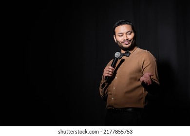 Skeptical Indian Stand Up Comedian Telling Joke Into Microphone On Dark Stage On Black