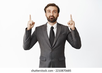 Skeptical Male Entrepreneur Pointing Fingers Up And Looking At Promo Offer, Smirk With Unamused, Disappointed Face Expression, Standing Over White Background