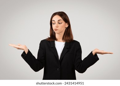 Skeptical millennial caucasian businesswoman in a tailored black suit with palms up in a balancing gesture, making a doubtful face as if weighing options or questioning a choice - Powered by Shutterstock