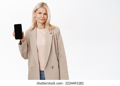 Skeptical Senior Woman, Stylish Grandmother Showing Mobile Phone Screen, Frowning Upset, Standing In Trench Coat Over White Background.