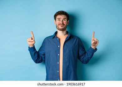 Skeptical Young Guy Smirk And Look Doubtful, Pointing Fingers Up Upset, Standing On Blue Background