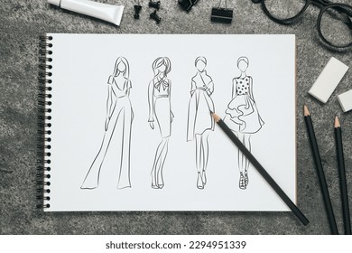 Sketches of different clothes in pad on grey textured table. Fashion designer's desk with stationery, flat lay