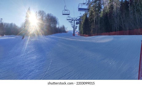 At The Ski Center, A Four-seat Chairlift Passes Directly Above The Ski Run. The Bright Sun Shines Through The Branches Of The Trees Along The Slope. Ski And Snowboard Tracks Are Visible In The Snow