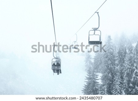 Ski lift above a snowy mountain with fir trees on a foggy day.