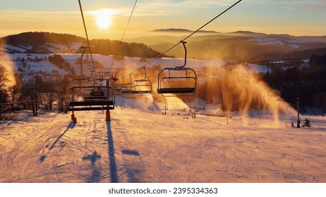 Ski lift empty ropeway on hilghland alpine mountain winter resort on bright sunny evening . Ski chairlift cable way with people enjoy skiing and snowboarding. Sunset sky backlit shining on background - Powered by Shutterstock