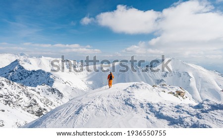 Ski mountaineer walking up along a steep snowy ridge with the skis in the backpack. Climber in a orange jacket climbs a mountain against a blue sky. Adventure concept. Panoramic view 