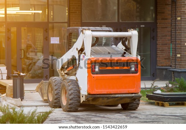 Skid loader stands in front\
of entrance to new multi-storey house and waits for garbage to be\
loaded into  bucket. Mini bulldozer waiting for cargo in front of\
porch