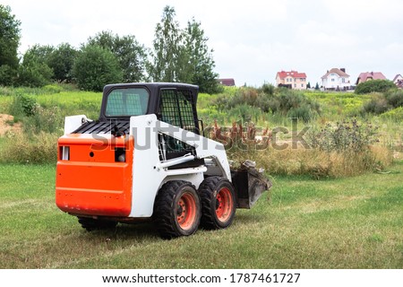 A skid steer loader clears the site for construction. Land work by the territory improvement. Machine for work in confined areas. Small tractor with a bucket for moving soil, turf and bulk materials.