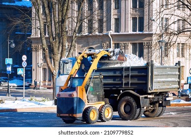 Skid Steer Loader Loading Pile Of Snow On Dump Truck, Snow Removing At City Street. Small Wheel Loader Clear Snow And Loading Dump Truck, Snow Hauling And Relocation Service. 