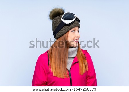 Skier redhead woman with snowboarding glasses over isolated blue wall looking to the side