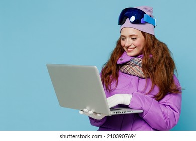 Skier smiling happy woman in warm purple padded windbreaker jacket ski goggles mask spend extreme weekend in mountains hold use work on laptop pc computer isolated on plain blue background studio.