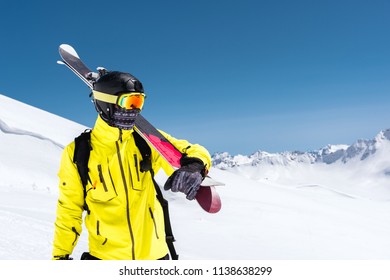 Skier Standing On A Slope. Man In A Light Suit, The Helmet And Mask In Skiing Is To Ski. In The Background Snow-capped Mountains , Skiers . Caucasus Mountains, Elbrus, Russia