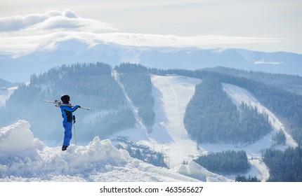 Skier standing on top of the mountain and enjoying the view on beautiful winter mountains on a sunny day. Woman is wearing blue ski suit, holding skis on her shoulder. Carpathian Mountains, Bukovel