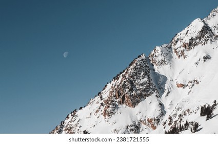 A skier stands atop a snow-covered mountain peak, illuminated by the glint of a crescent moon in the twilight sky - Powered by Shutterstock