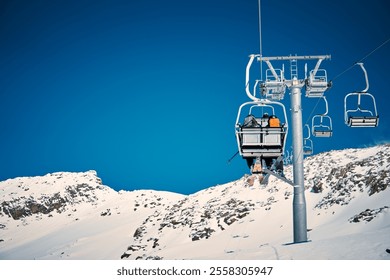 Skiers enjoying a scenic ride on a ski lift against a clear blue sky in a snowy mountain landscape - Powered by Shutterstock