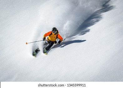 A skiier making a turn in deep snow and sunshine creating a large ploom of snow and a shadow