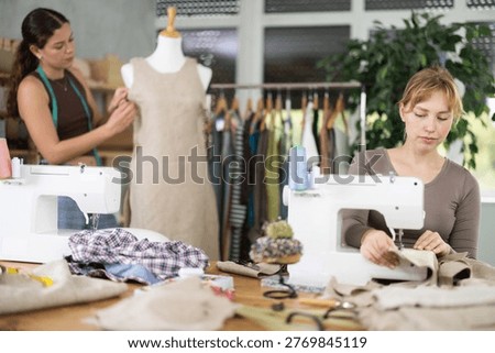 Skilful young tailoress using sewing-machine while other working with mannequin in sewing workshop