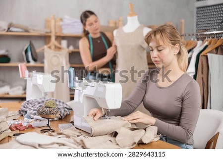 Skilful young tailoress using sewing-machine while other working with mannequin in sewing workshop