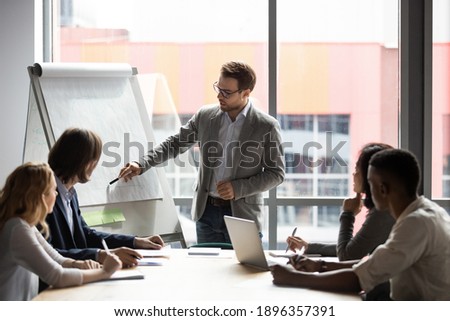 Skilled 30s caucasian leader in eyeglasses coaching interested business people, giving educational workshop presentation in office. Concentrated employees listening to professional coach at workshop.