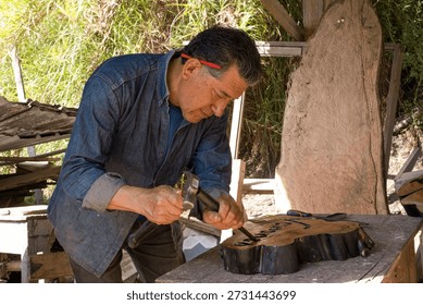 Skilled artisan man using a chisel and hammer to carefully carve intricate letters and patterns into a tree stump board. Creating a unique piece of handcrafted woodwork in an outdoor workshop setting