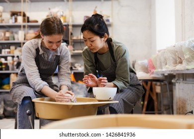 Skilled Asian Potter In Apron Teaching Young Woman To Make Clay Vase On Pottery Wheel In Modern Workshop, Pottery Course