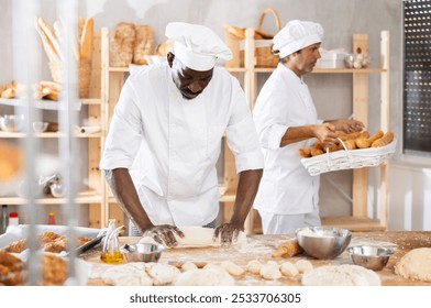 Skilled black baker expertly rolling dough into long loaf shape, preparing for baking in warm rustic bakery environment, surrounded by freshly baked bread and pastries arranged on wooden shelves - Powered by Shutterstock