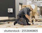 Skilled carpenter assembles wooden panels in a factory, using tools like an electric screwdriver and drill to build a house framework. Dressed in protective gear, he showcases his craftsmanship