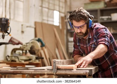 Skilled carpenter cutting a piece of wood in his woodwork workshop, using a circular saw, and wearing safety googles and earmuffs, with other machinery in the background