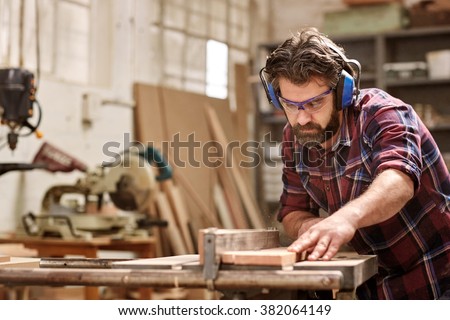 Skilled carpenter cutting a piece of wood in his woodwork workshop, using a circular saw, and wearing safety googles and earmuffs, with other machinery in the background
