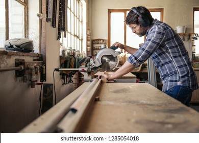 Skilled carpenter wearing safety gear sawing a piece of wood with a mitre saw while working alone in his woodworking studio - Powered by Shutterstock