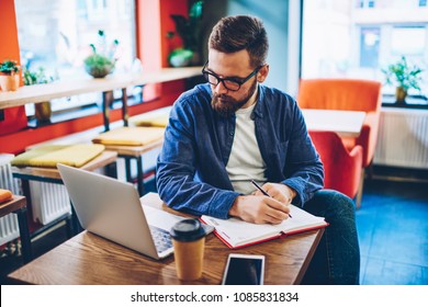 Skilled casual dressed student watching webinar online on digital netbook and making notes in notepad.Pensive bearded blogger rewriting information with creative ideas from website using 4G internet