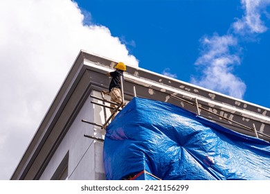 A skilled construction worker in a hard hat standing on top of scaffolding sanding the eaves and fascia of a 3 story house, in preparation for painting. Exterior renovation of a building facade.