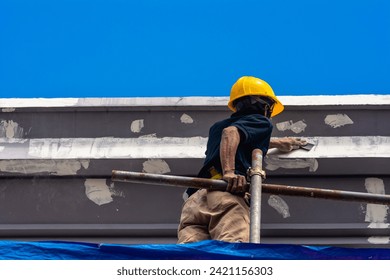 A skilled construction worker in a hard hat standing on top of scaffolding sanding the eaves and fascia of a 3 story house, in preparation for painting. Exterior renovation of a building facade.